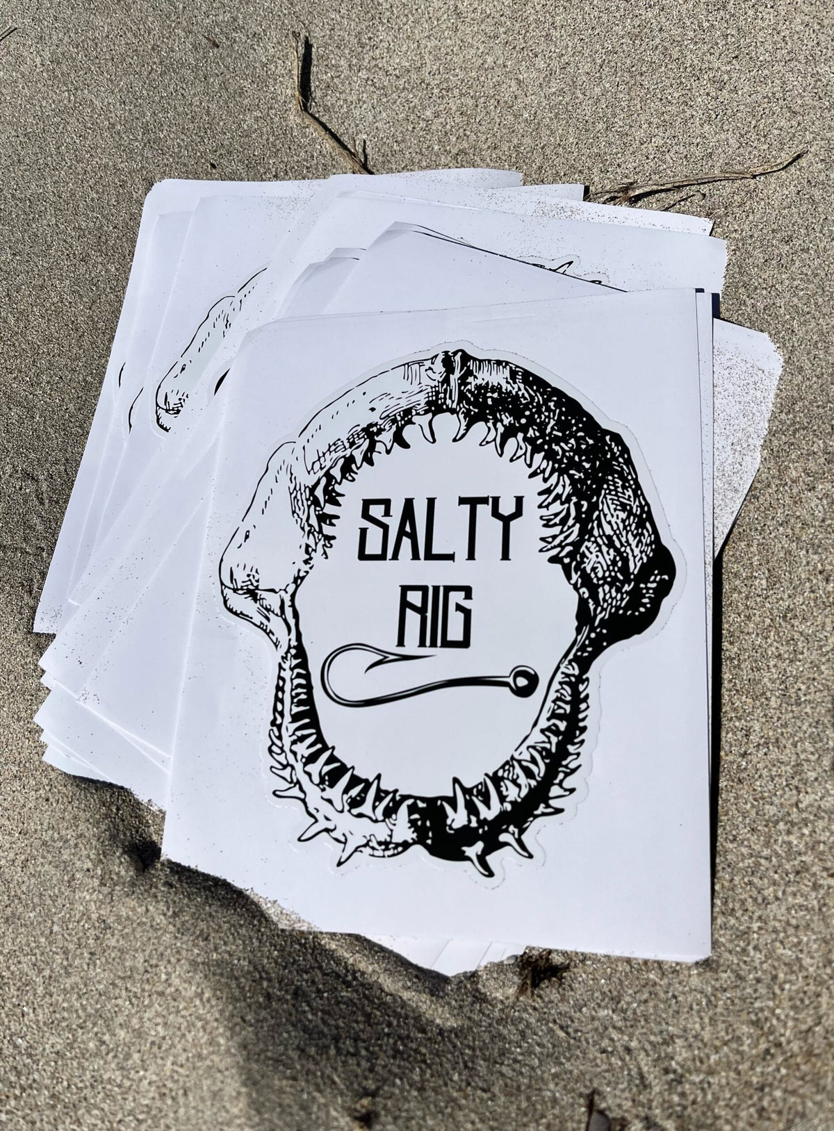 Stack of prints with a shark tooth design and 'Salty Rig' text on a sandy surface