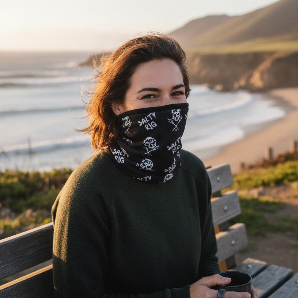 A woman sitting on a wooden bench outdoors by the ocean, wearing a black neck gaiter with a white 'Salty Pig' graphic print. The background shows a scenic coastal landscape with cliffs, beach, and waves during golden hour.