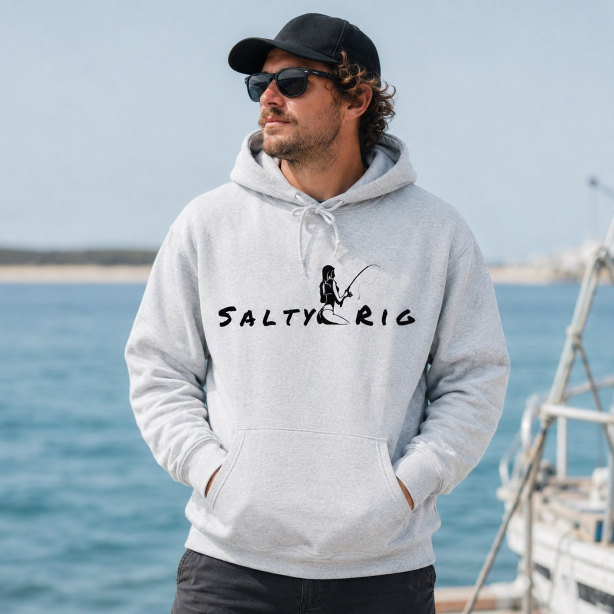 A man wearing a light gray heather hoodie with a black 'SALTY RIG' fishing graphic stands by the ocean, wearing a black cap and sunglasses.